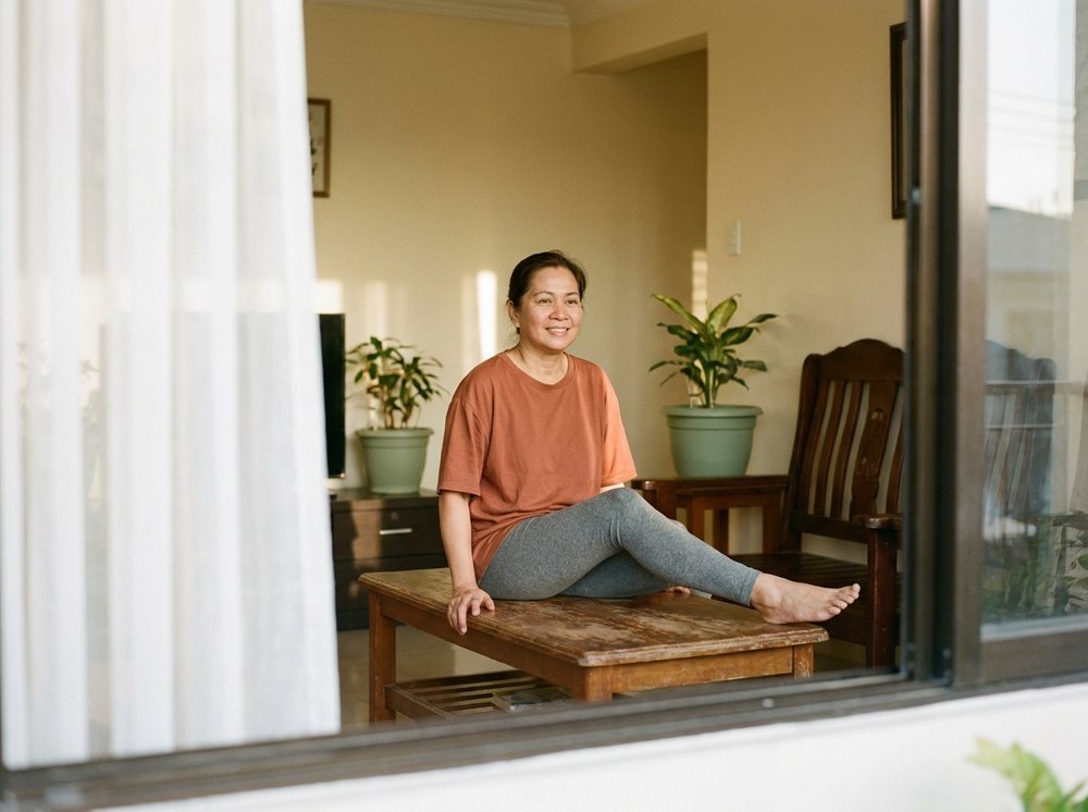 A Filipino woman in her sixties gently straightening her knee on a woven mat at home